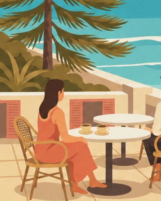 A woman in a coral dress seated at a table with 2 cups of coffee, facing the sea
